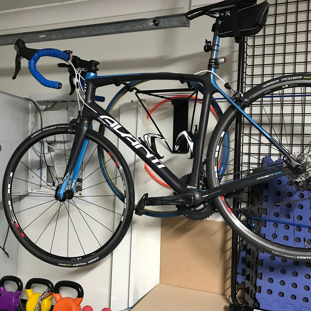 Black and blue road bike with drop handlebars hanging on a wall rack in a garage above colorful kettlebells.
