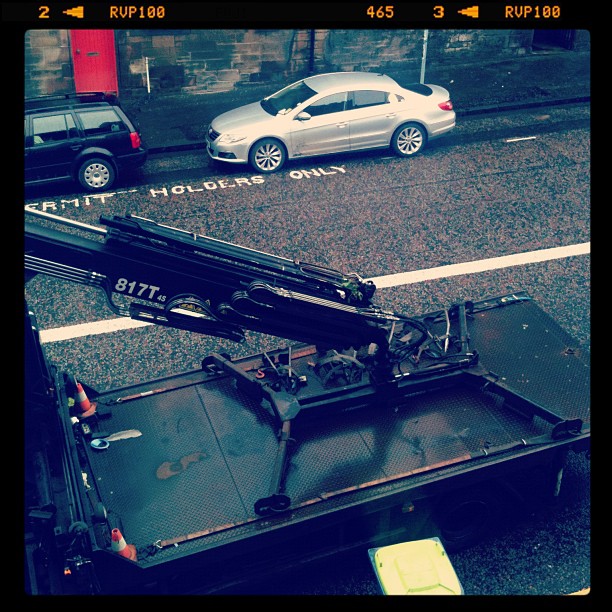 Tow truck bed with folded crane parked on a street beside two cars and road markings reading “permit holders only”.