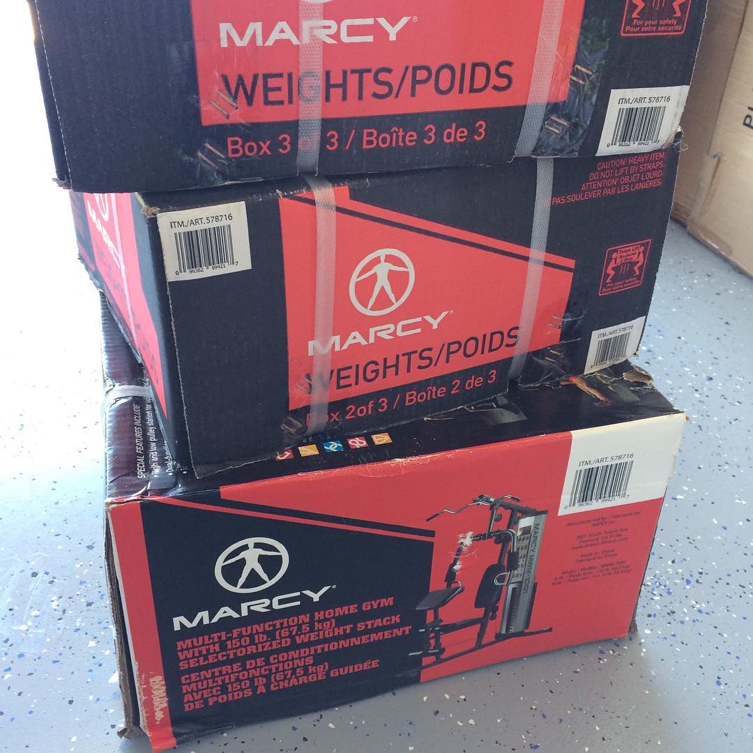 Three stacked Marcy boxes labeled weights and home gym sit on a speckled floor, secured with plastic straps.