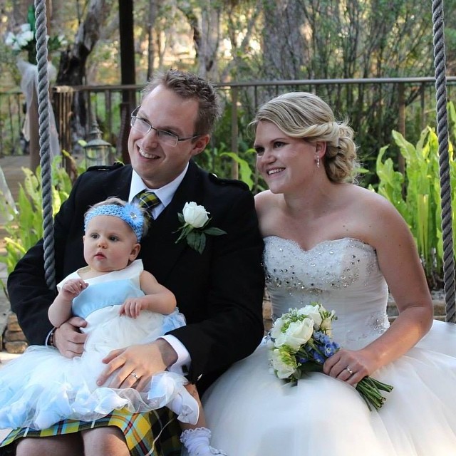 Bride and groom in wedding attire sit smiling with a baby in a blue and white dress on a rope swing outdoors.