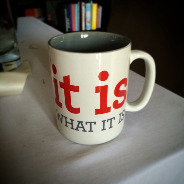 White mug with a gray interior and red and black text, sitting on a table near a glass and books in the background.