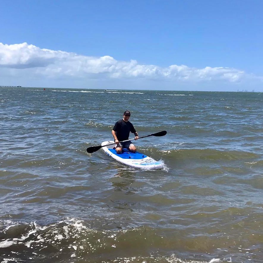 Person kneeling on a blue and white paddleboard in choppy ocean water under a bright blue sky with low clouds.