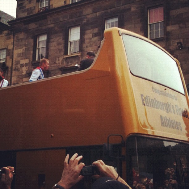 Open-top double-decker bus with people on the upper deck, viewed from below, with hands holding cameras in the foreground.