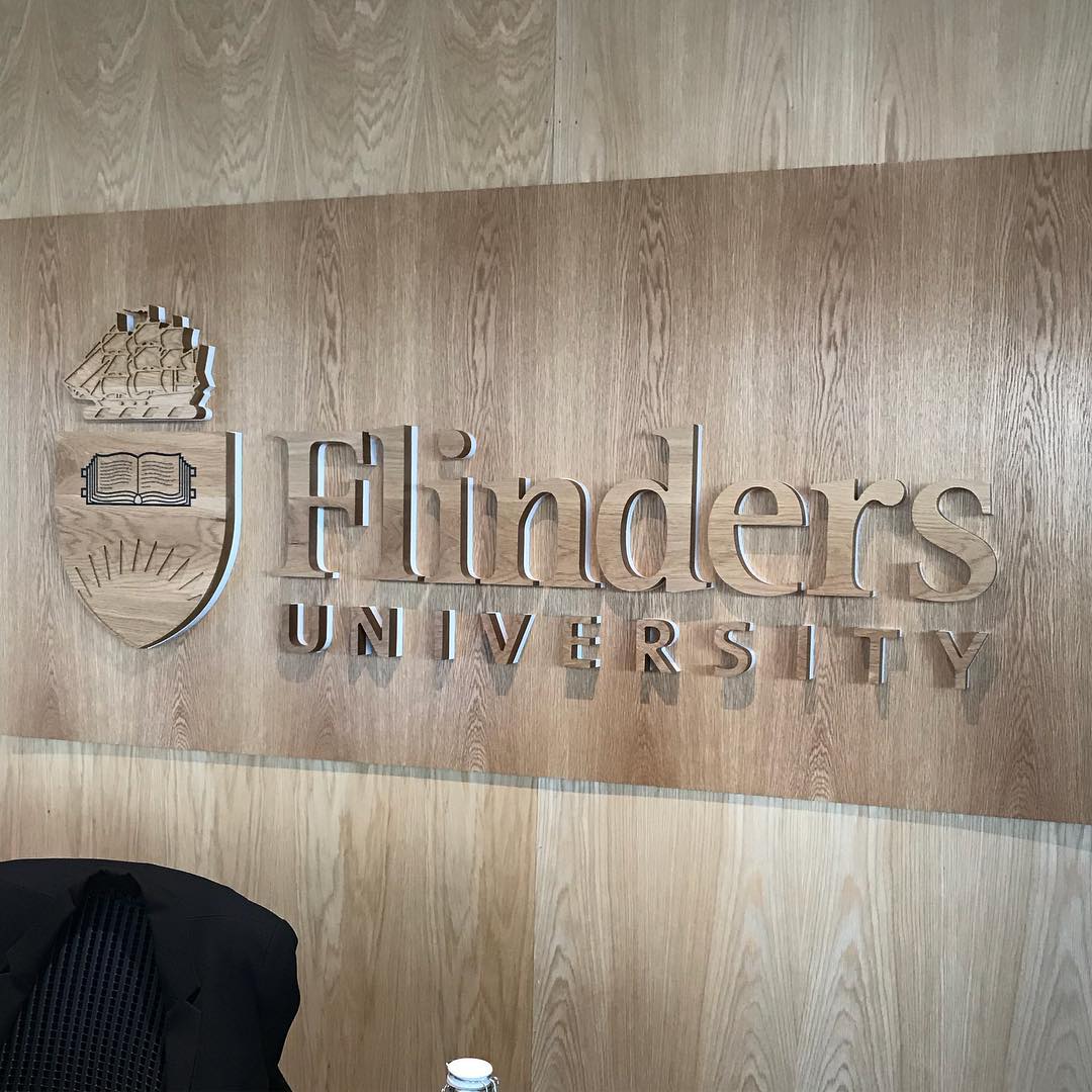 Flinders University sign and crest mounted on a wood-paneled wall, with part of a dark chair visible in the lower left.