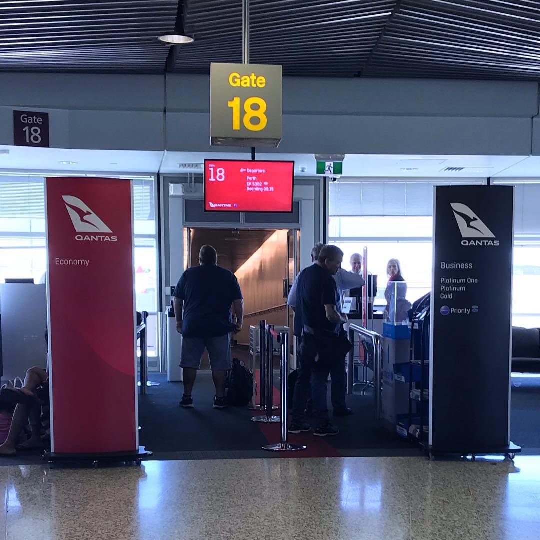Airport gate 18 boarding area with Qantas signs for economy and business, a departure screen, and several people near the entrance.