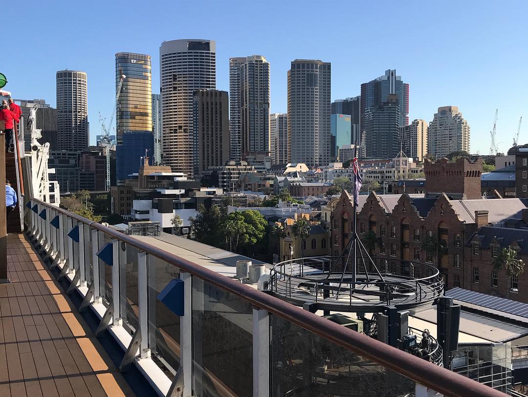 City skyline with tall modern towers and older brick buildings, viewed from a ship deck with glass railing under a clear blue sky.