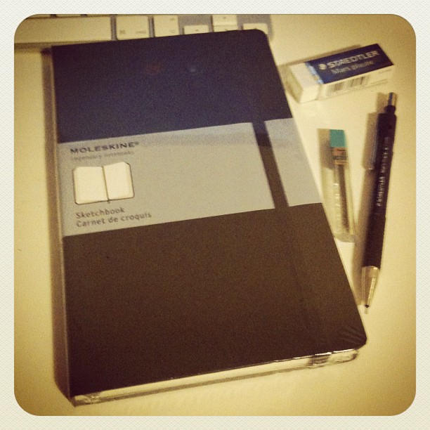 Black Moleskine sketchbook on a desk beside a mechanical pencil, lead refill tube, and Staedtler eraser.