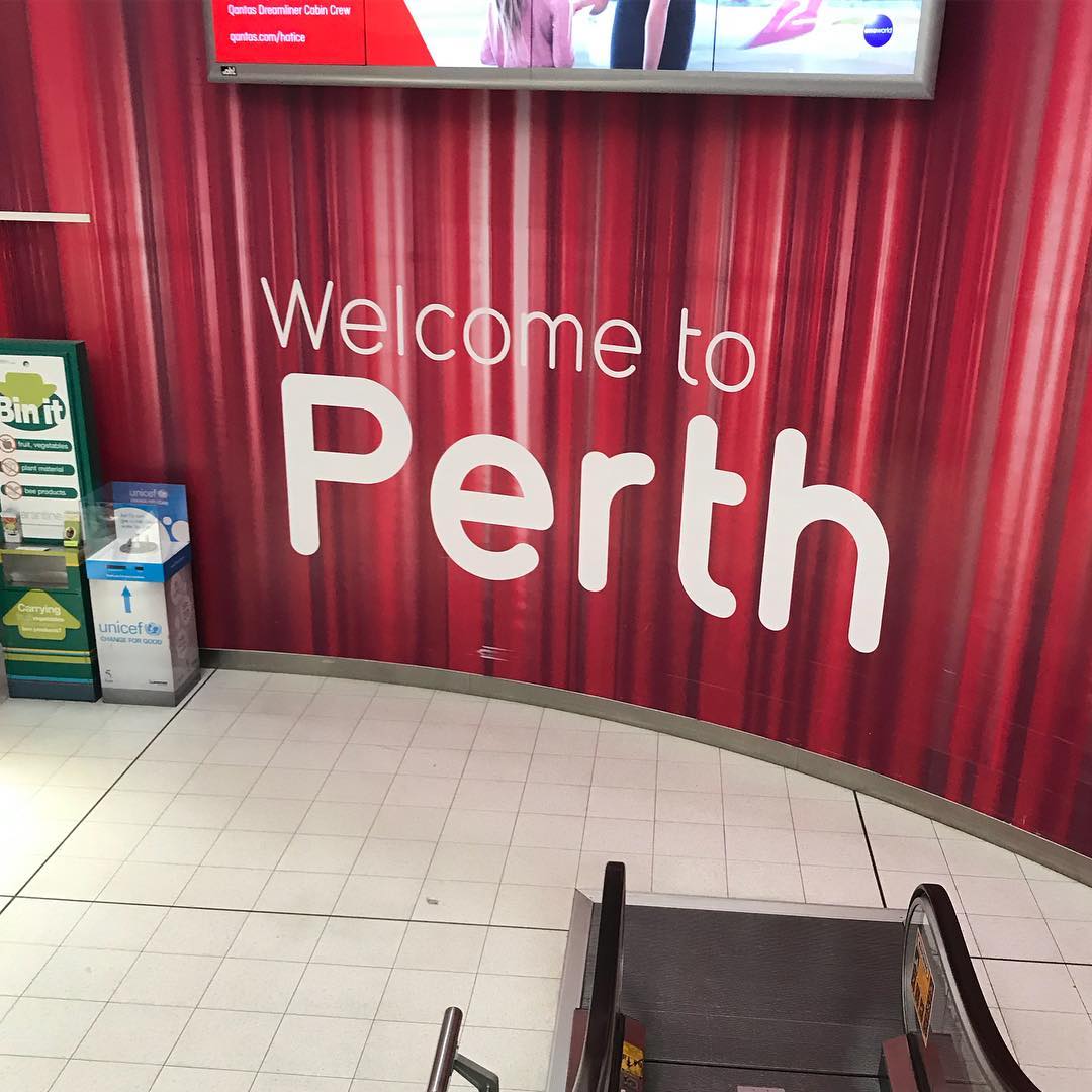Red wall with large white text reading "Welcome to Perth" beside recycling bins and the top of an escalator.