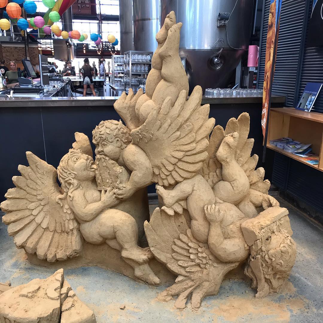 Intricate sand sculpture of winged cherubs and figures displayed inside a brewery or bar with colorful lanterns overhead.