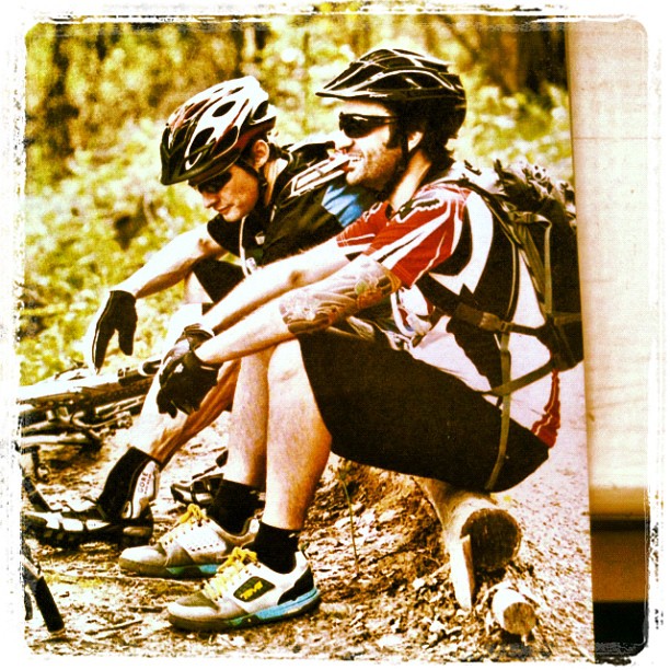 Two cyclists wearing helmets and gloves sit resting on a log beside their bikes in a wooded area.