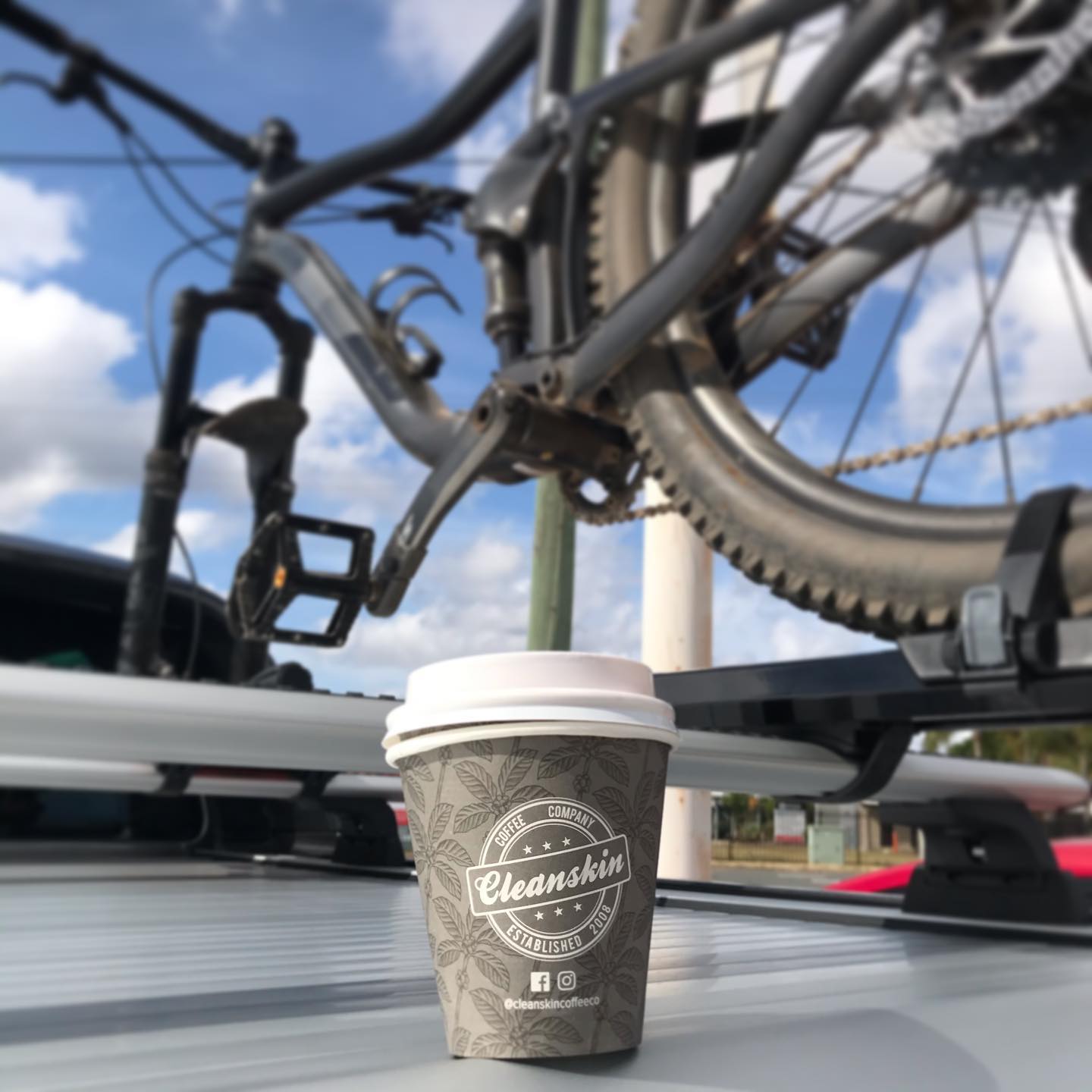 Coffee cup on a car roof rack beneath a mounted bicycle, with a blue sky and clouds in the background.