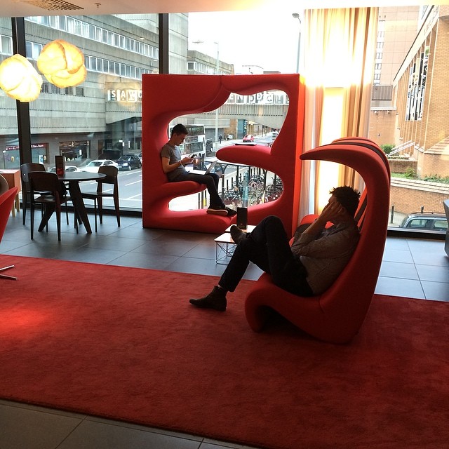 Two people sit in sculptural red chairs near large windows in a modern lounge with tables, street views, and a red carpet.