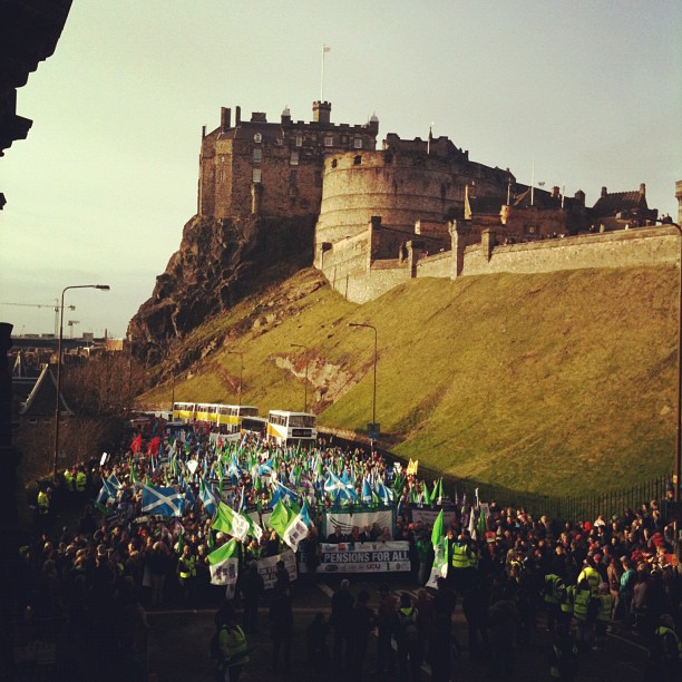 Large crowd carrying blue, green, and white flags marches below a hilltop stone castle.