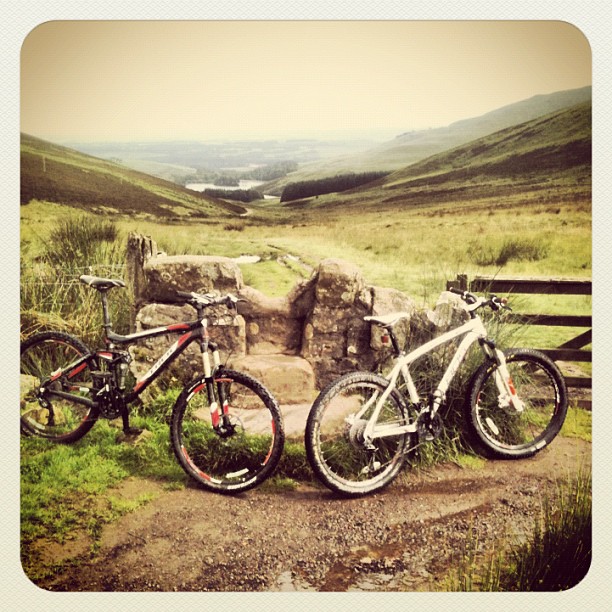 Two mountain bikes beside a stone stile and wooden gate in a grassy valley with rolling hills in the distance.