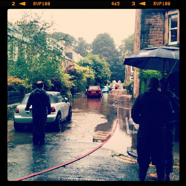 Rain-soaked residential street with floodwater, parked cars, a person walking, and another holding an umbrella near a brick building.