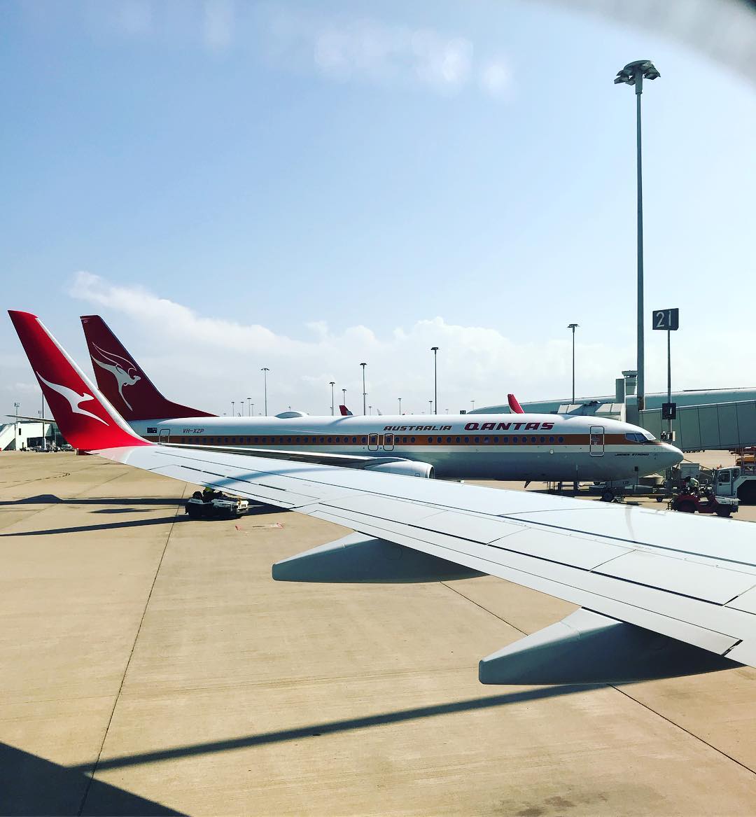 View from an airplane window of a wing on the tarmac beside a Qantas jet at gate 21 under a clear blue sky.
