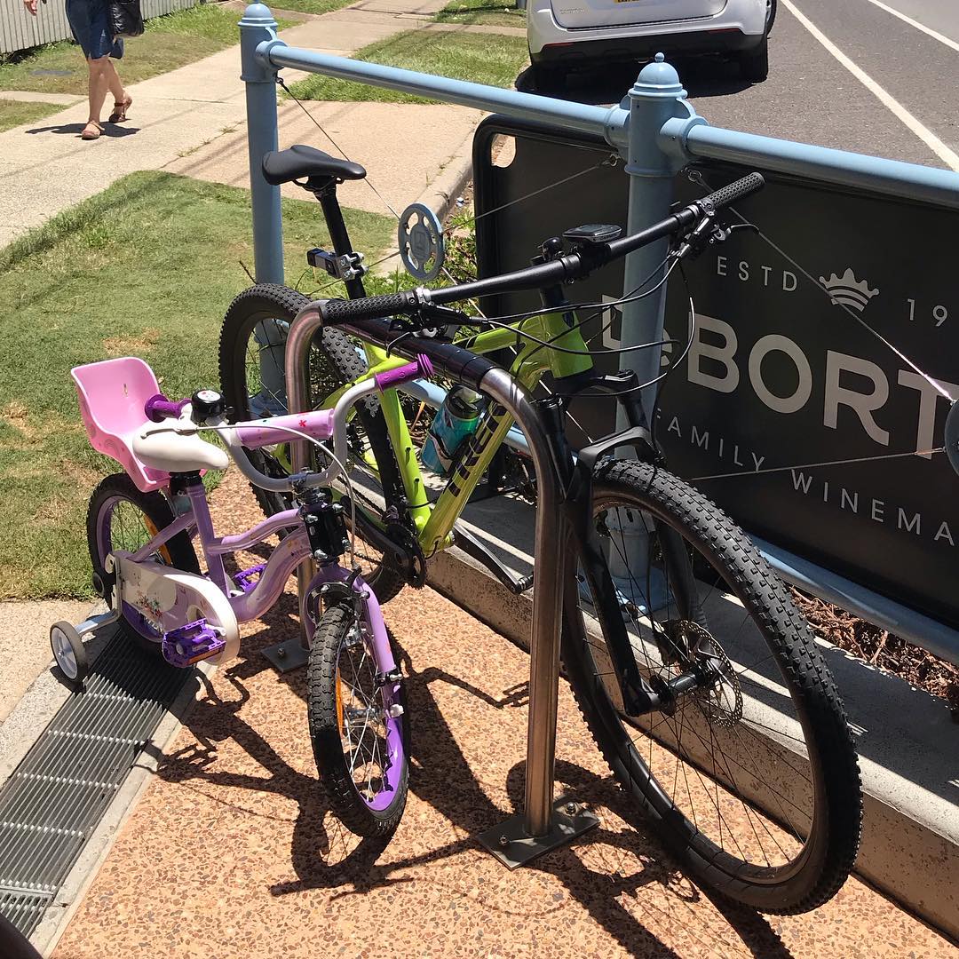 A pink child’s bike and a green mountain bike are locked to a sidewalk bike rack beside a street.