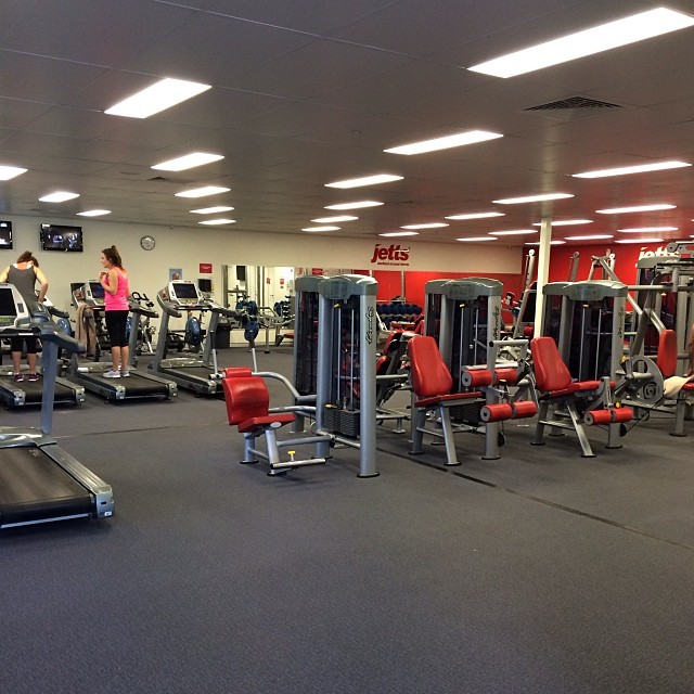 Gym interior with treadmills, red and gray weight machines, mirrors, and two people exercising.