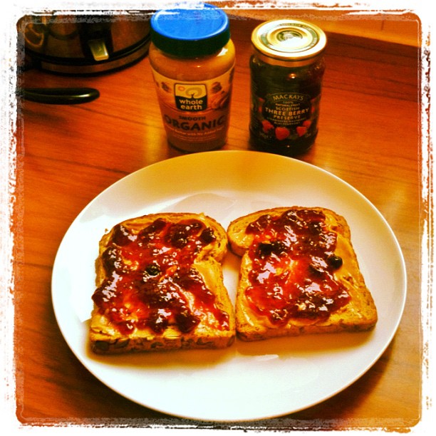 Two slices of toast with peanut butter and berry jam on a white plate, with peanut butter and jam jars on a wooden table.
