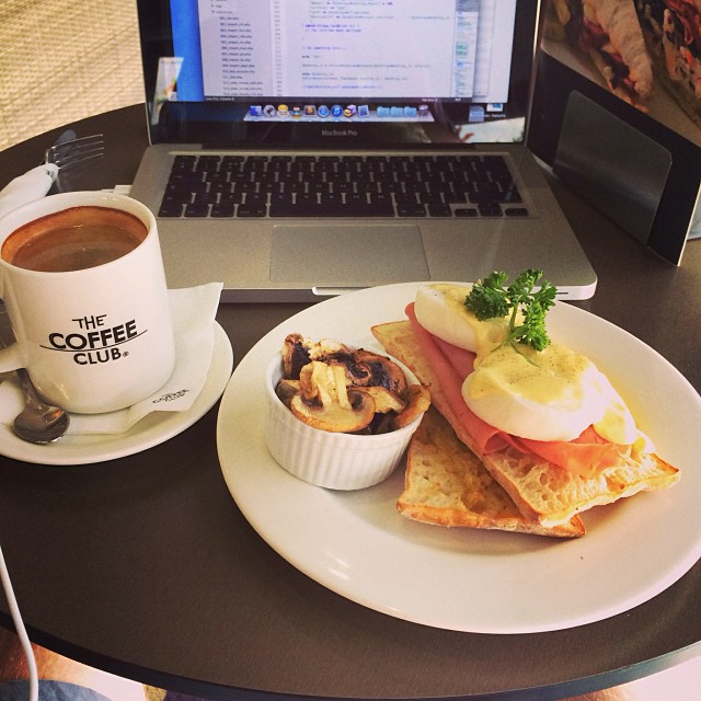 Coffee beside a laptop and a plate with eggs Benedict on toast and a small dish of mushrooms on a café table.