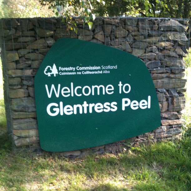 Green sign reading "Welcome to Glentress Peel" in front of a wire stone wall with grass and trees around it.