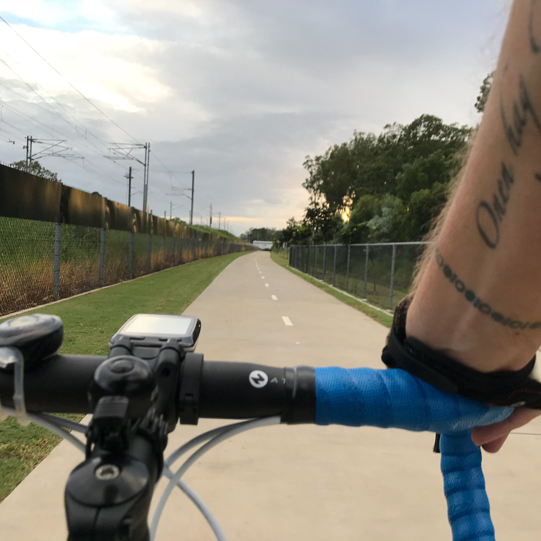 View from bicycle handlebars on a paved path between fences, with a tattooed arm on blue-wrapped bars under a cloudy sky.