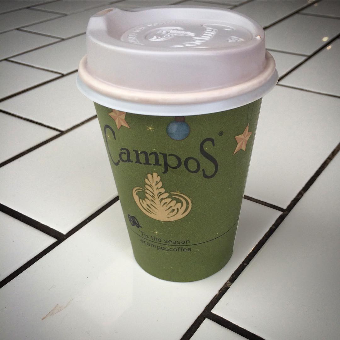 Green Campos coffee cup with a white plastic lid on a white tiled surface.
