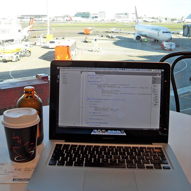 Laptop displaying code on a table beside a coffee cup and bottle, with airport runways and parked airplanes visible through the window.