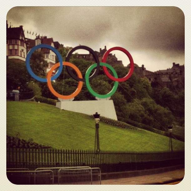 Large Olympic rings sculpture on a grassy hillside, with old buildings and cloudy sky in the background.