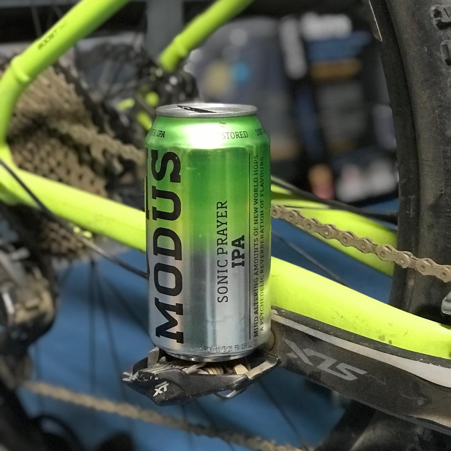 Green Modus Sonic Prayer IPA can balanced on a bicycle pedal beside the chain and rear wheel.