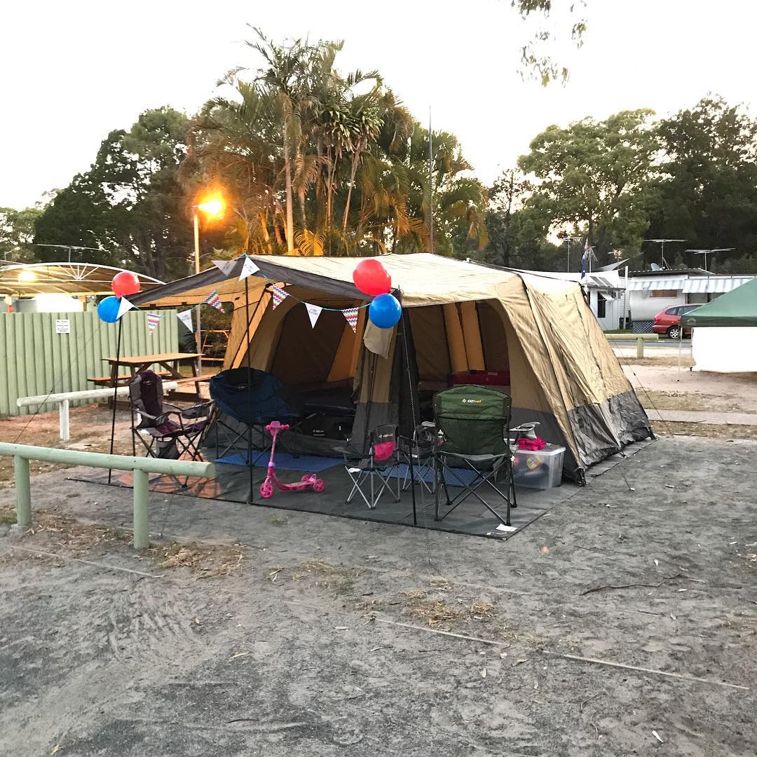 Large tan camping tent decorated with bunting and red and blue balloons, surrounded by folding chairs and a pink scooter.