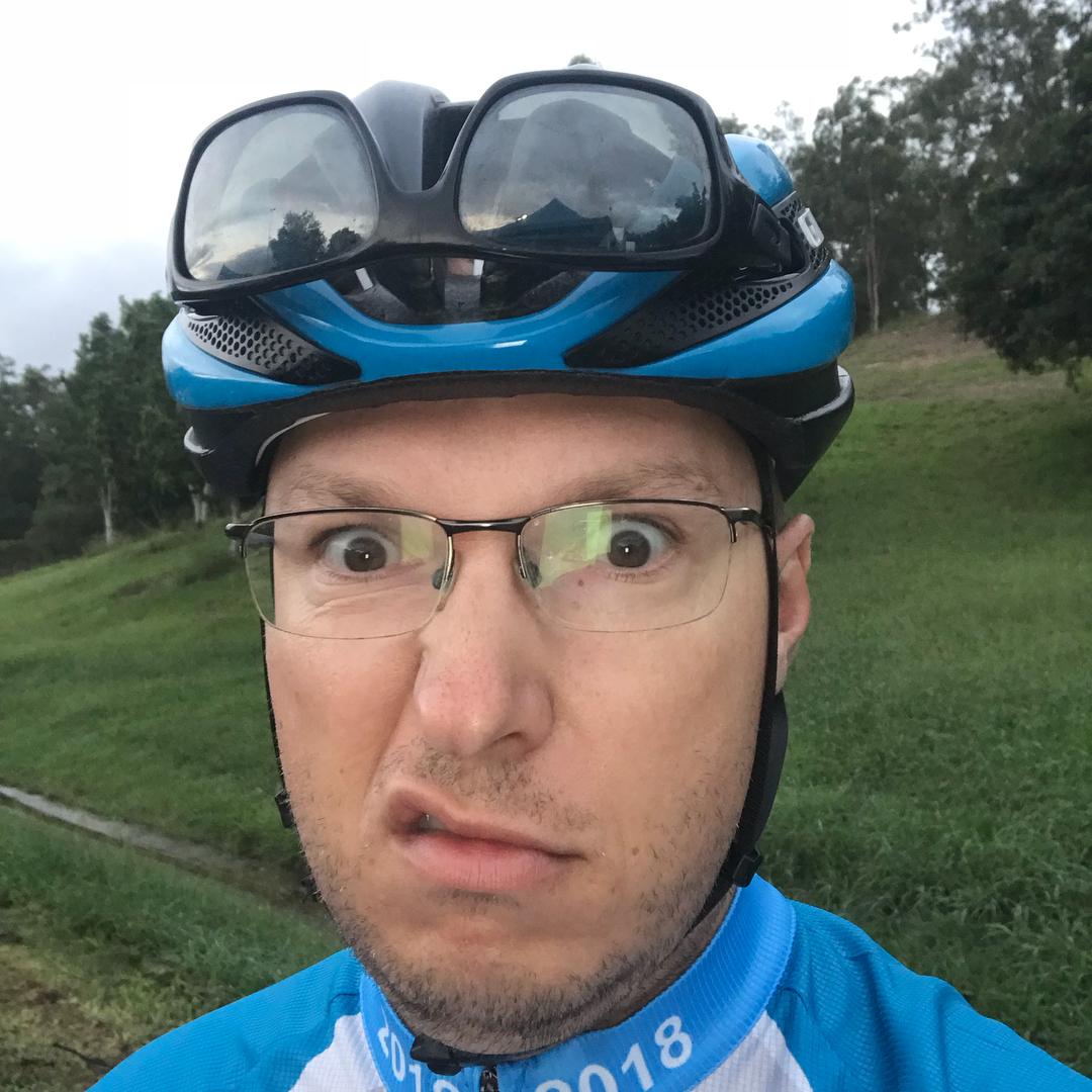 Person wearing glasses and a blue bike helmet with sunglasses on top, making a confused face outdoors on a grassy path.