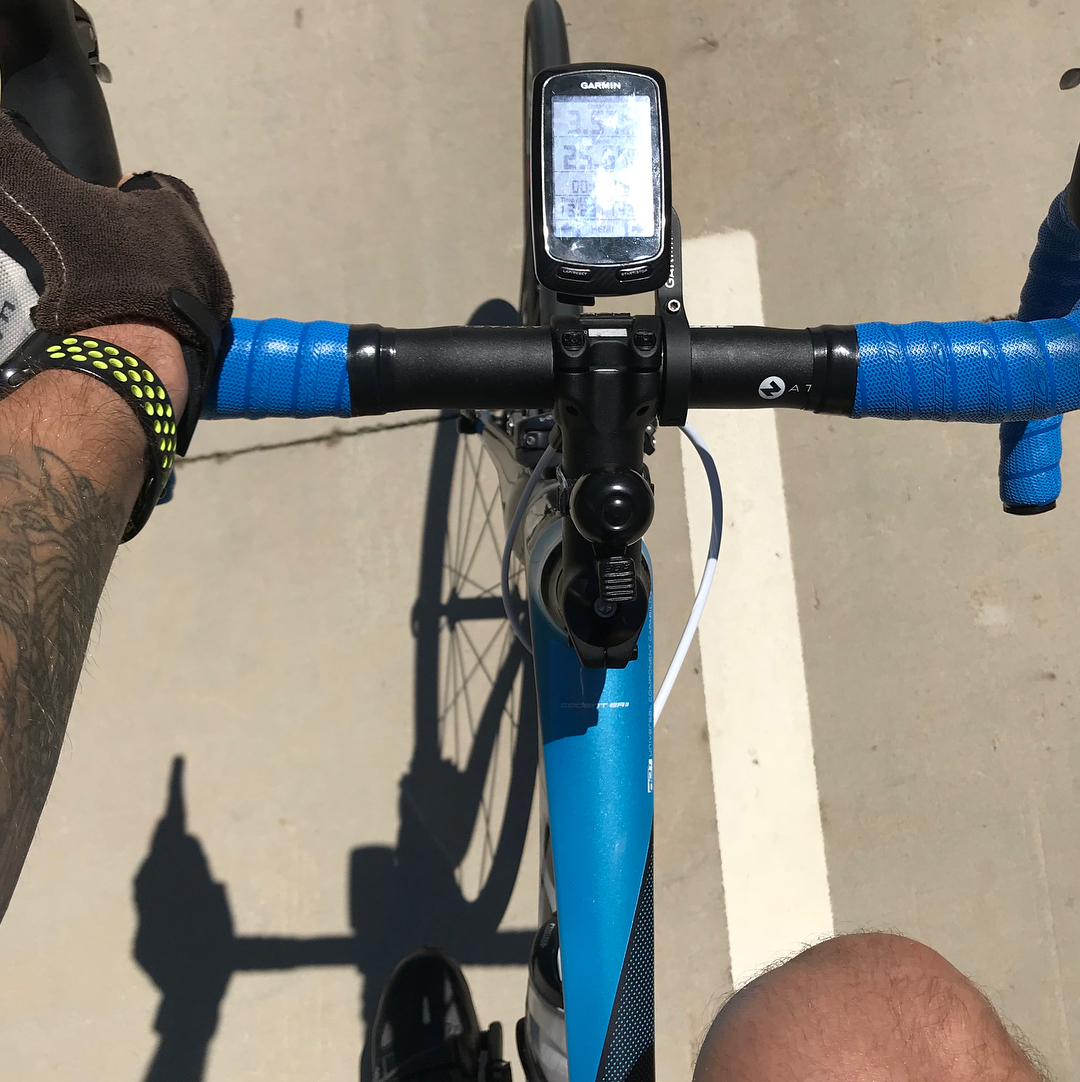 View from a cyclist riding a road bike, with blue handlebar tape, a Garmin computer, and gloved hands on a sunlit paved road.