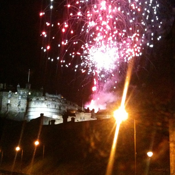 Fireworks burst in pink and white above a lit stone castle at night, with streetlights glowing in the foreground.