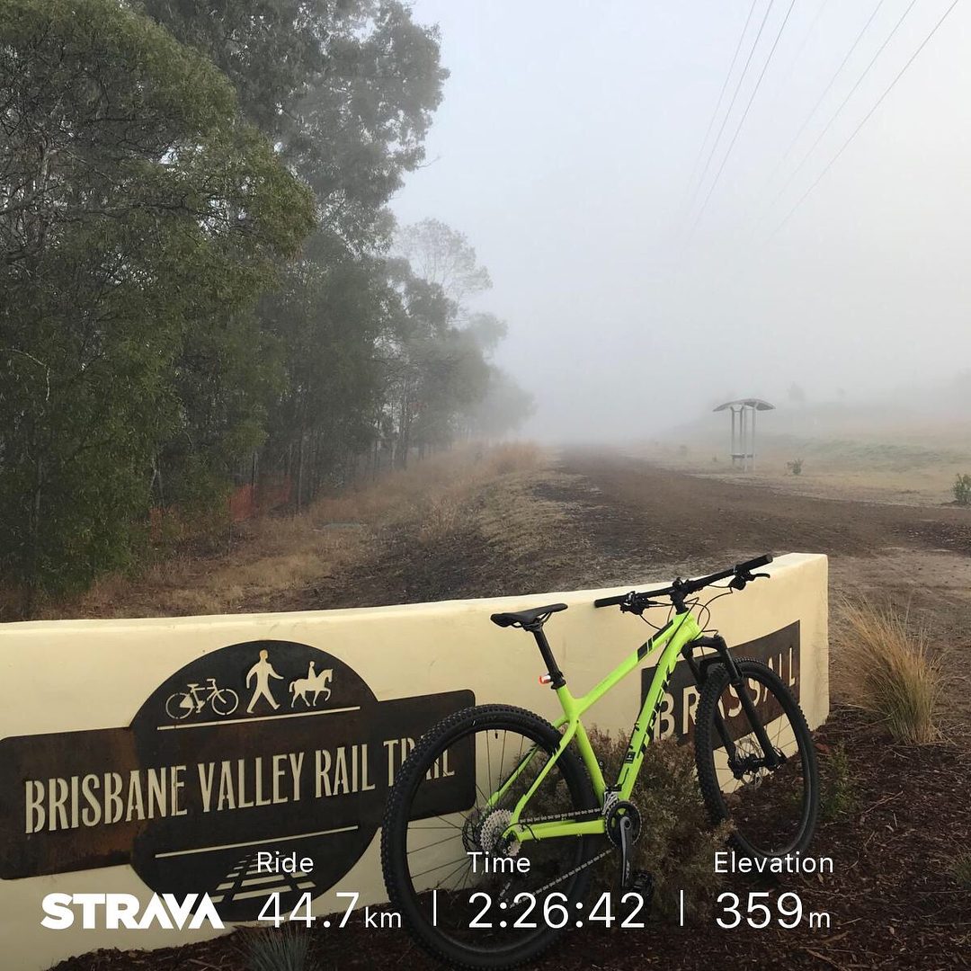 Green mountain bike leaning against a Brisbane Valley Rail Trail sign beside a foggy dirt path lined with trees.