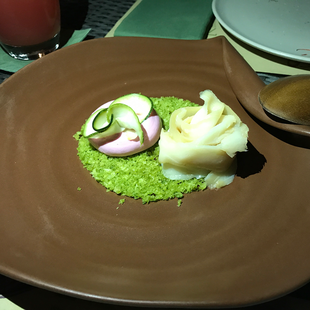 Plated dessert with a pink dome and white rose-shaped garnish on bright green crumbs in a large brown bowl.
