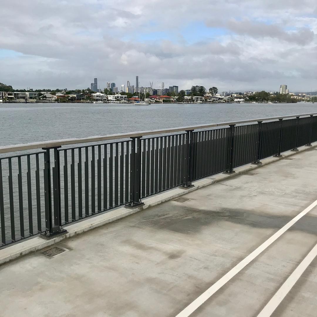 Concrete waterfront walkway with black railing, calm river, low houses and sailboats across the water, and a distant city skyline under cloudy sky.