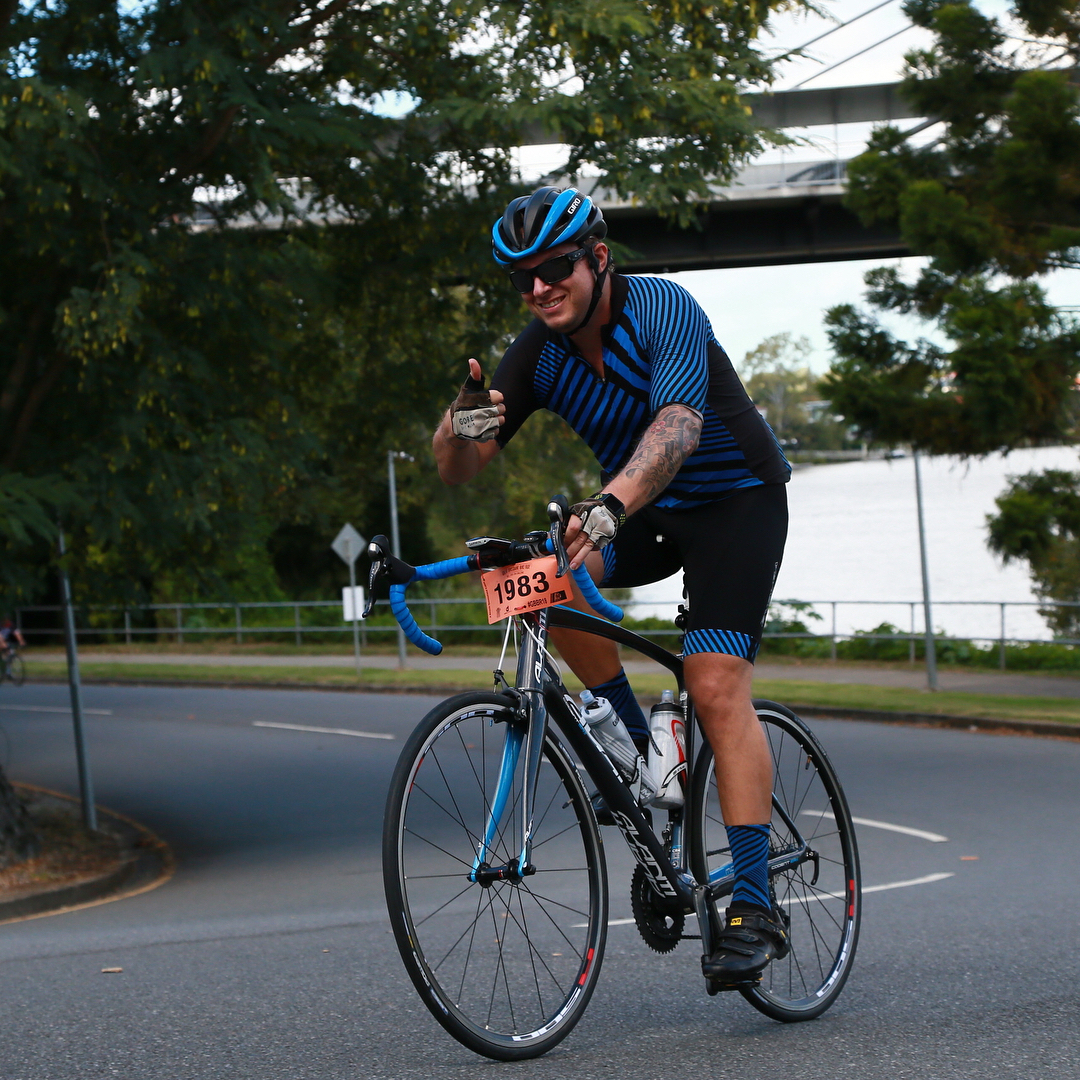 Cyclist in a blue-and-black kit rides a road bike on a tree-lined street, giving a thumbs-up, with a bridge and water behind.