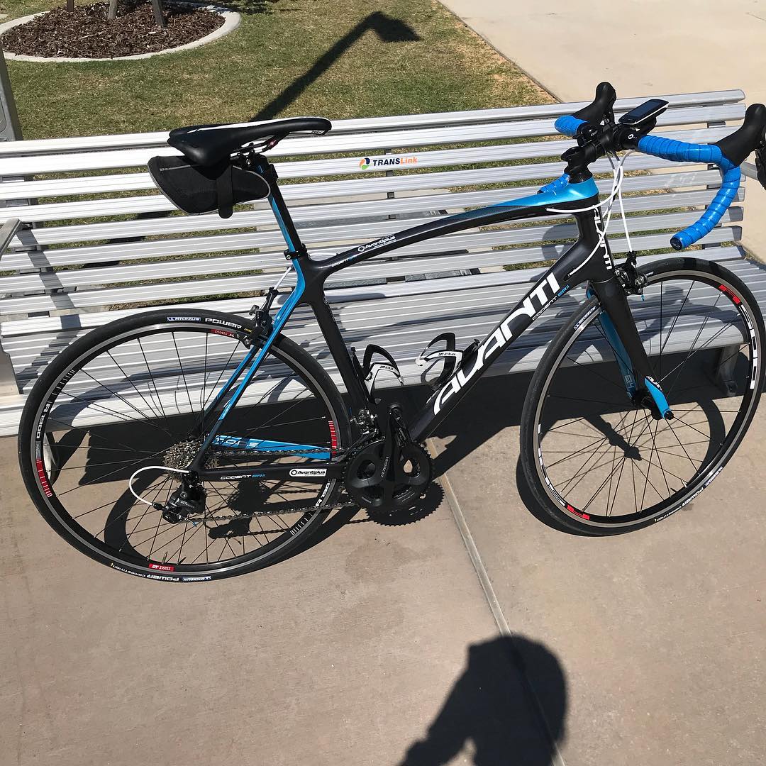 Black and blue road bike with drop handlebars leaning against a silver bench in bright sunlight.