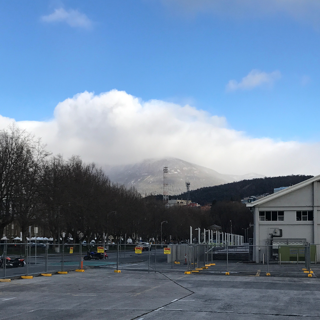 Clouds rolling over a mountain behind leafless trees, a fenced parking lot, and a white building under a bright blue sky.