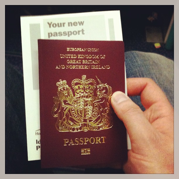 Hand holding a maroon UK passport over a paper leaflet titled "Your new passport" on a lap.