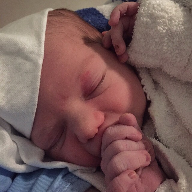 Sleeping newborn in a white cap and blanket with tiny hands clasped near their mouth.