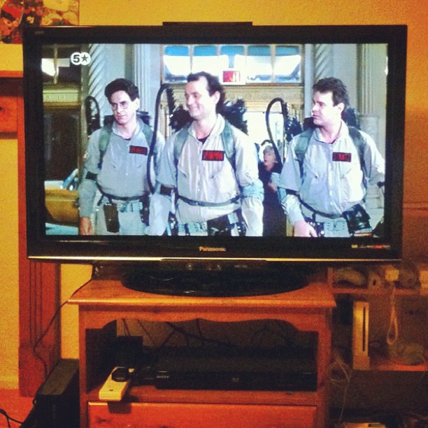 Television on a wooden stand showing three men in gray jumpsuits with backpack gear walking indoors.