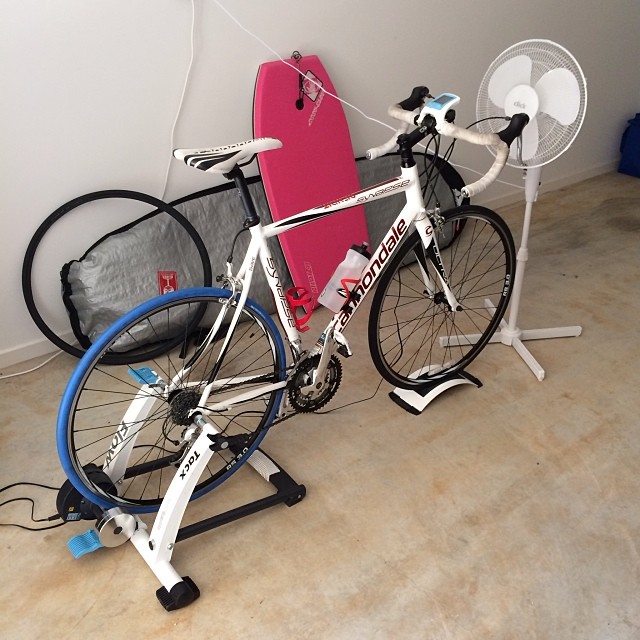 White road bike on an indoor trainer with a pink bodyboard behind it, a standing fan beside it, and spare wheels leaning against the wall.