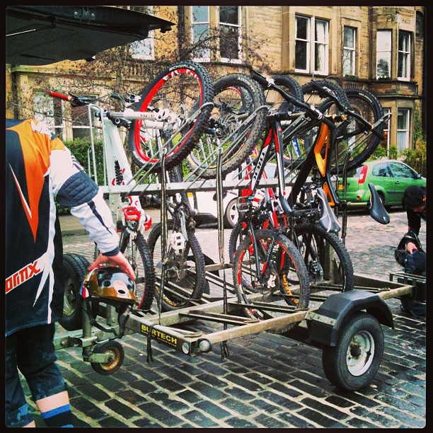 Several mountain bikes secured upright on a small trailer, with a person in cycling gear holding a helmet beside it.