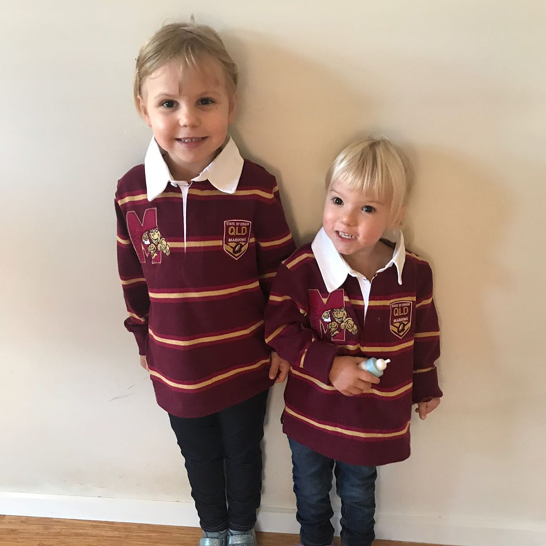 Two young children in matching maroon striped rugby shirts stand against a beige wall, with the younger child holding a small bottle.