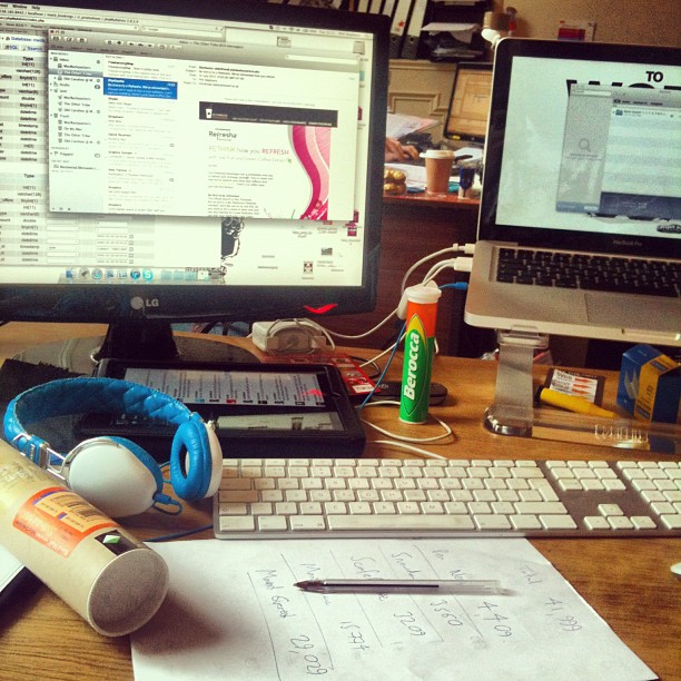 Desk with two computer monitors, a keyboard, blue headphones, a notepad, a pen, and a green tube on a cluttered workspace.