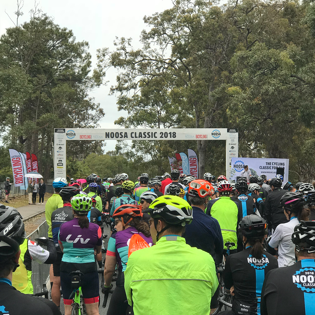 Cyclists in helmets and bright jerseys gather beneath a “Noosa Classic 2018” start banner on a tree-lined road.