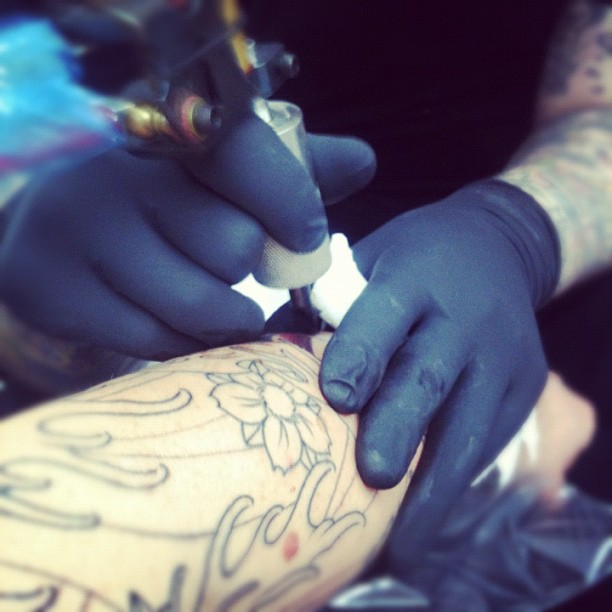 Blue-gloved hands use a tattoo machine on a person's forearm with floral and script outlines.