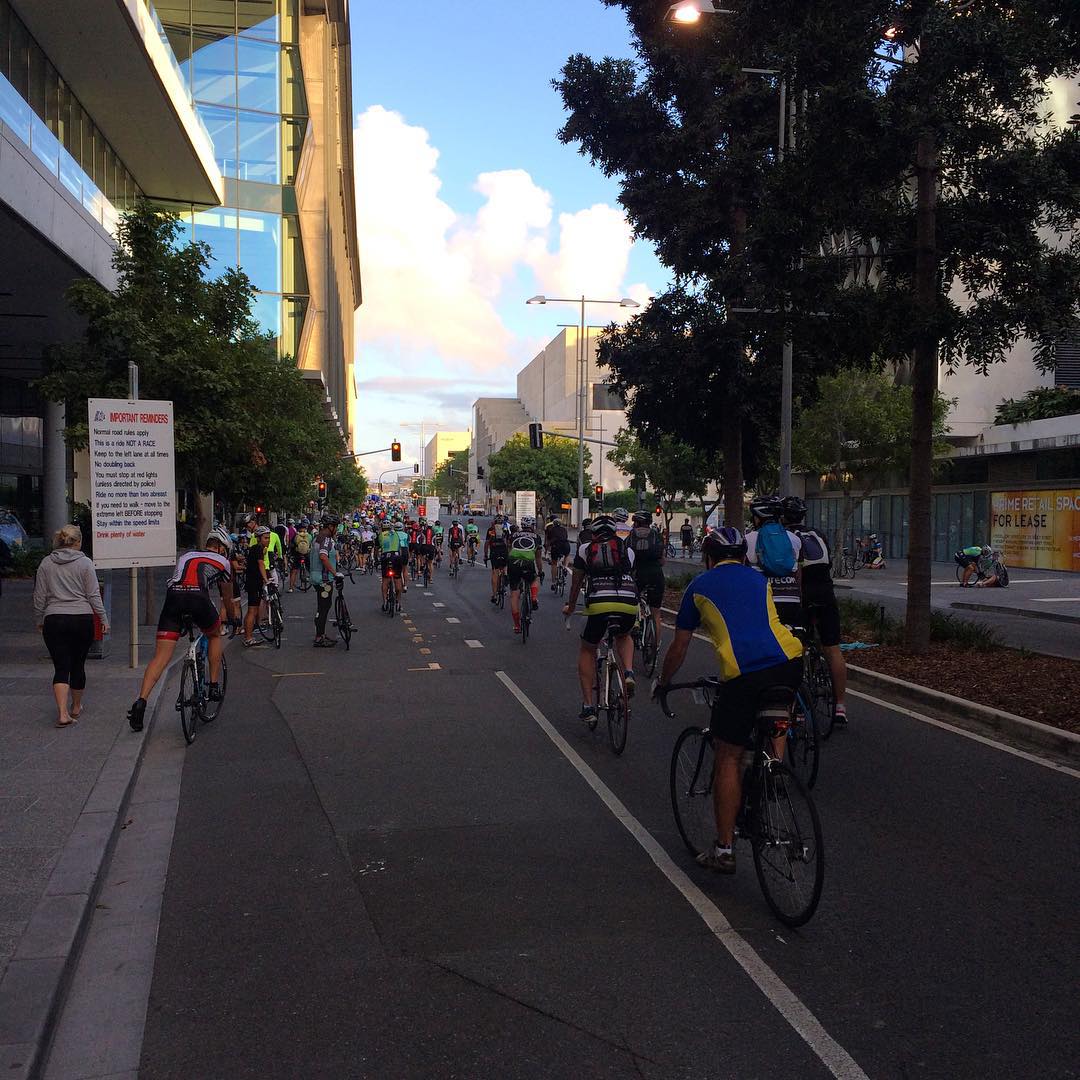 Dozens of cyclists ride down a city street between tall buildings and trees under a bright cloudy sky.